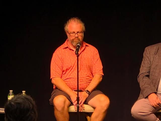 A bearded man with glasses, in an orange polo shirt and shorts, sits quietly on a stool at a microphone on a dark stage.