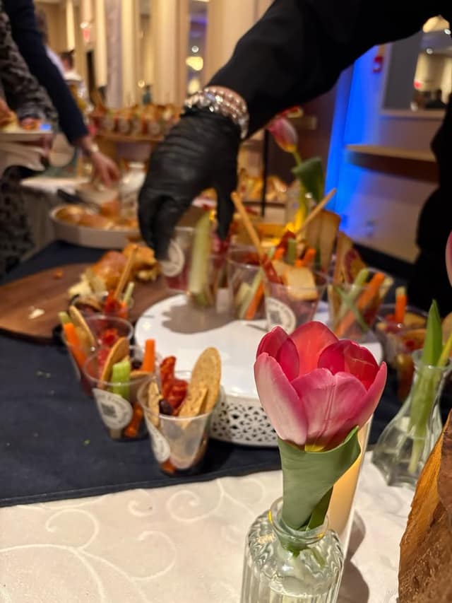 A gloved hand reaches for snack cups of vegetables, crackers and cured meats beside a pink tulip in a glass vase.