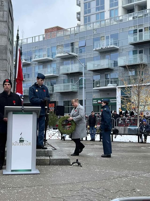 A woman in a gray coat carries a wreath past a podium marked Kitchener, as a cadet and a man in a poppy toque stand nearby.