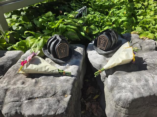 Two bronze firefighter helmet markers on stone blocks, labelled Kitchener Fire Fighter, each with a bouquet of flowers laid beside it.