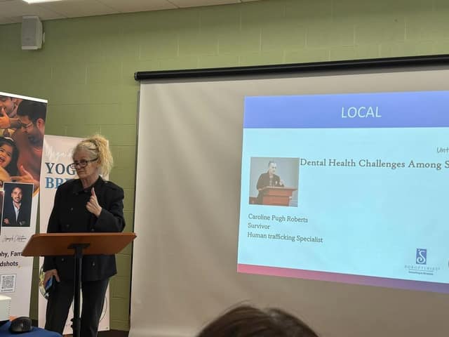 A woman in glasses and a dark jacket stands at a podium beside a projected slide reading "LOCAL" with Caroline Pugh Roberts listed.