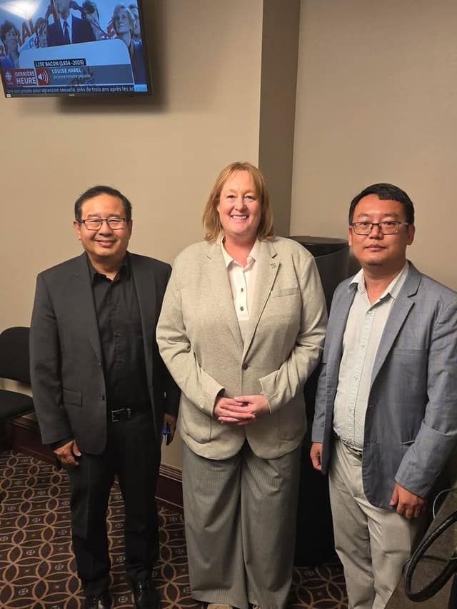 A woman in a beige blazer stands smiling between two men in glasses and blazers in a meeting room, with a TV news screen on the wall behind.