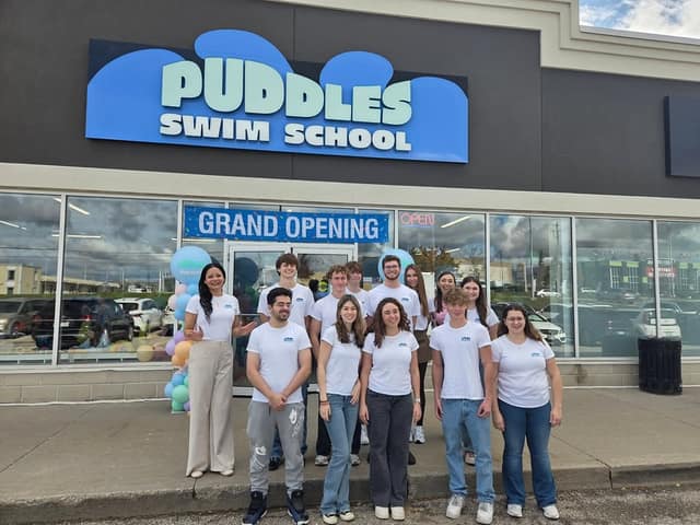 A group of about a dozen people in matching white shirts stands outside Puddles Swim School under a Grand Opening banner and balloons.