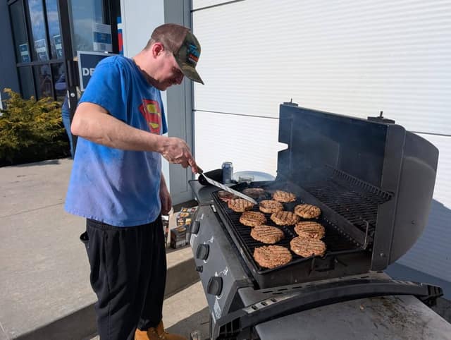 A man in a camouflage cap and blue Superman T-shirt flips burgers on a barbecue grill outside a building.