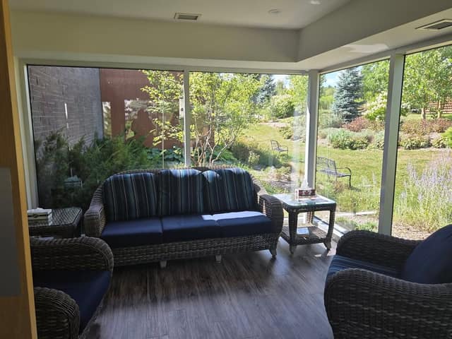 A sunroom with a wicker sofa and armchairs with navy cushions, overlooking a garden with benches, shrubs and trees.