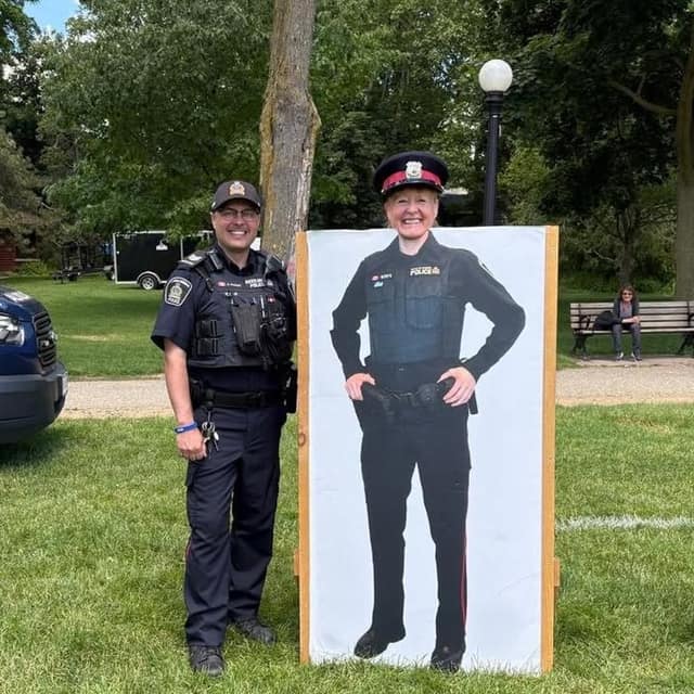 A smiling police officer in uniform stands beside a life-size cardboard cutout of a uniformed officer in a park.