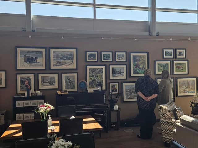 Two women stand with their backs to the camera, viewing framed landscape paintings on a wall above a piano and a dining table.