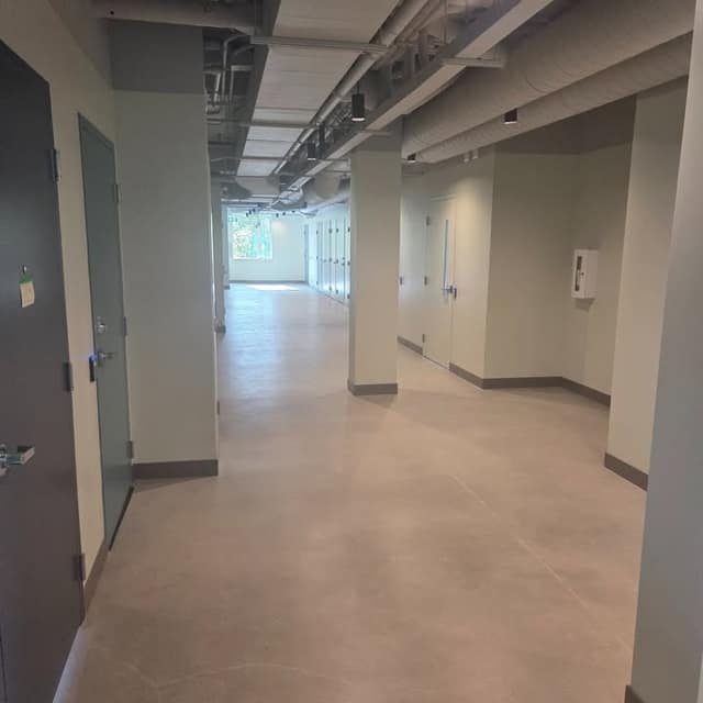 A long concrete-floored hallway with gray doors on the left, exposed ducts overhead, and a window at the far end.
