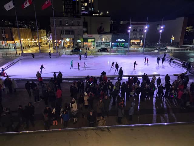 A crowd in winter clothing gathers at night beside a lit outdoor skating rink where skaters glide, with flags and downtown buildings behind.