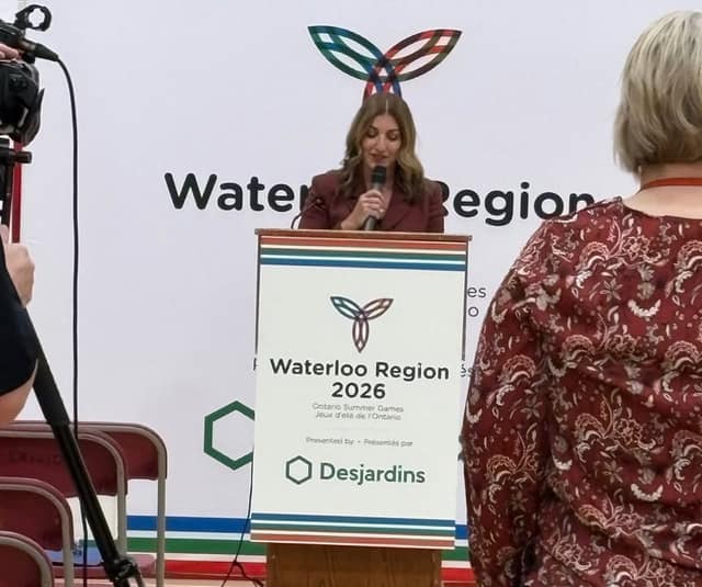 A woman in a burgundy blazer speaks into a microphone at a podium reading Waterloo Region 2026, while a camera films the event.