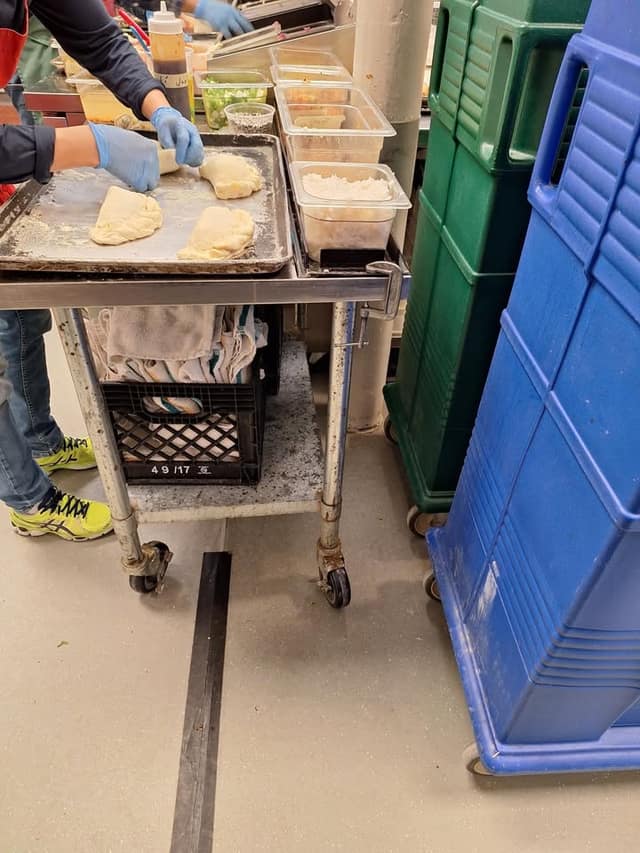 A volunteer in blue gloves shapes dough on a baking tray atop a metal cart in a kitchen, beside food tubs and large bins.