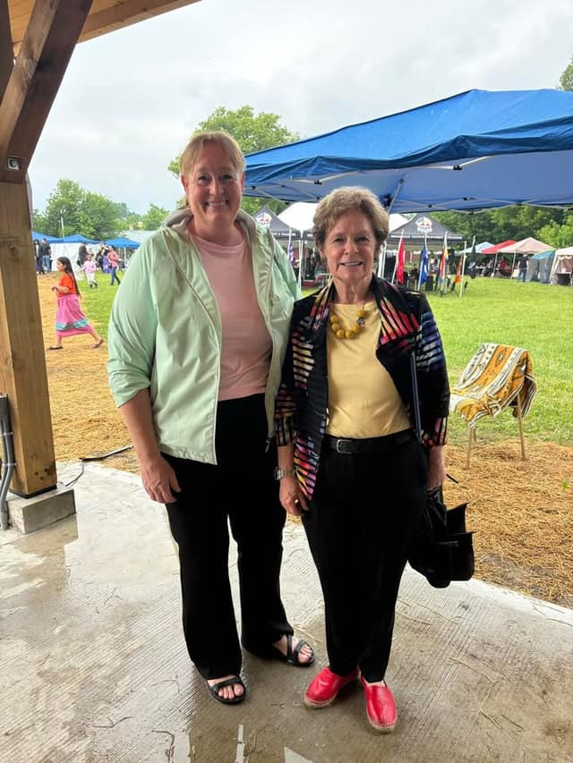 Two smiling women stand under a covered wooden structure, with vendor tents, flags and a grassy powwow ground behind them.