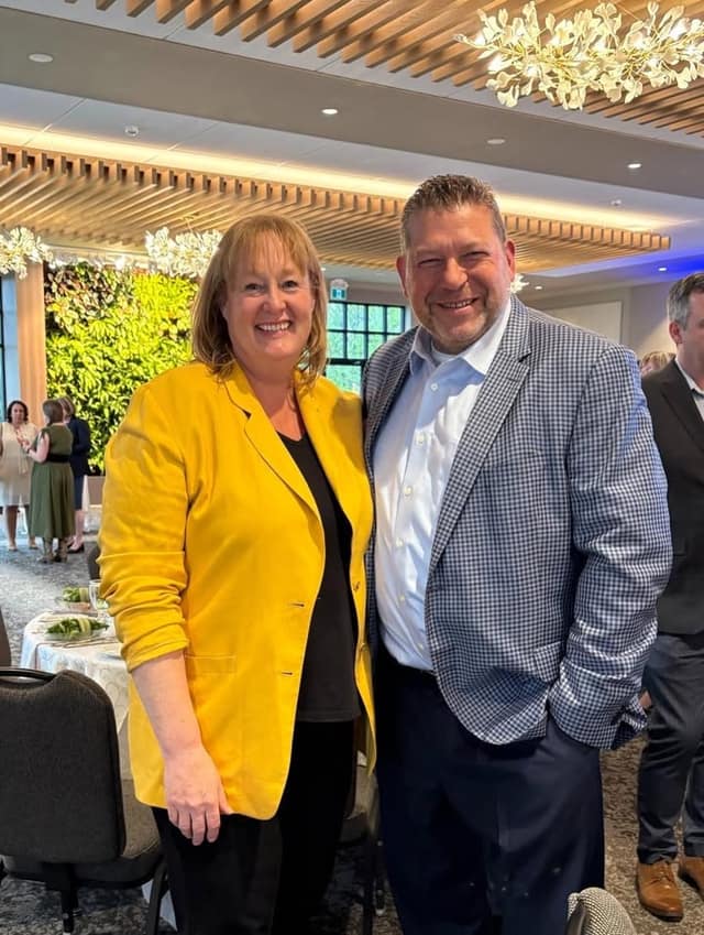 A smiling woman in a yellow blazer stands beside a smiling man in a blue checked blazer in a banquet hall with a green plant wall.