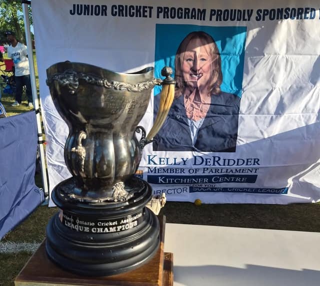 A silver trophy marked League Champions sits before a banner reading Junior Cricket Program Proudly Sponsored, with Kelly DeRidder's portrait.