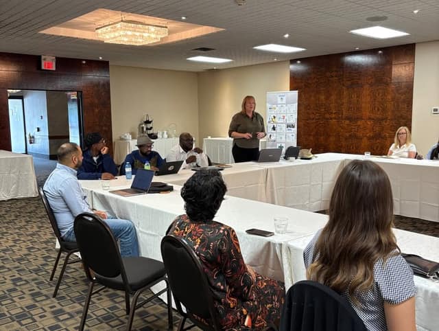 A woman stands speaking to attendees seated at white-draped tables with laptops in a banquet hall with a chandelier.