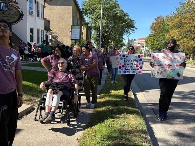 Walkers in purple Walk to Wellness shirts proceed along a sidewalk past a seniors' residence, with a resident in a wheelchair and a man holding a sign.