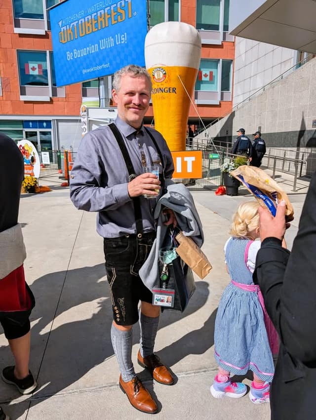 A man in lederhosen holds a glass and a bag beside a giant inflatable beer glass and an Oktoberfest banner; a girl stands nearby.