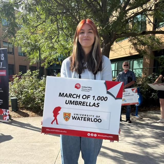 A smiling woman holds a sign reading "March of 1,000 Umbrellas" with United Way and University of Waterloo logos.