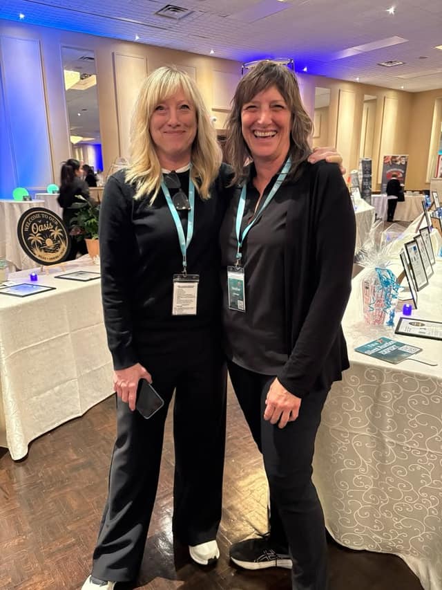 Two smiling women in black outfits wear lanyards and pose together in a ballroom beside tables of silent auction items.