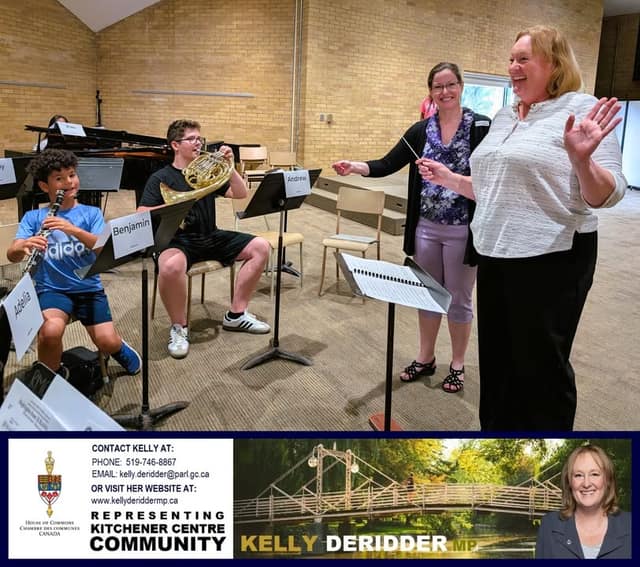A laughing woman waves a baton beside a smiling woman as a boy plays clarinet and another plays French horn at music stands in a band room.