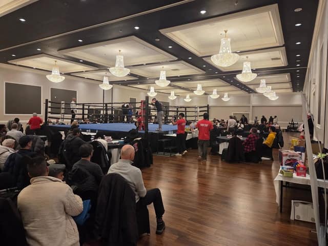 A ballroom with chandeliers, a boxing ring and seated spectators, with event staff in red shirts standing near tables.
