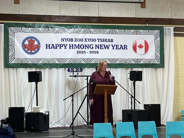 A woman in a maroon dress speaks into a microphone at a wooden lectern beneath a banner reading "Happy Hmong New Year 2025-2026."