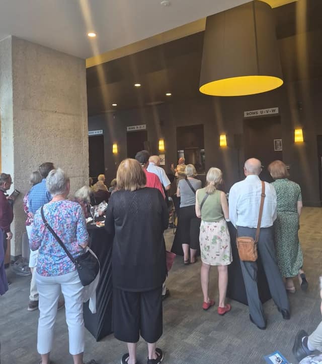 Attendees stand in a theatre lobby facing a speaker at the back, under a large yellow lamp shade and signs marking seating rows.