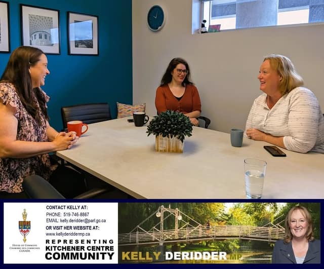 Three women laugh and talk around a white table with mugs and a plant in a meeting room with a teal wall and a clock.