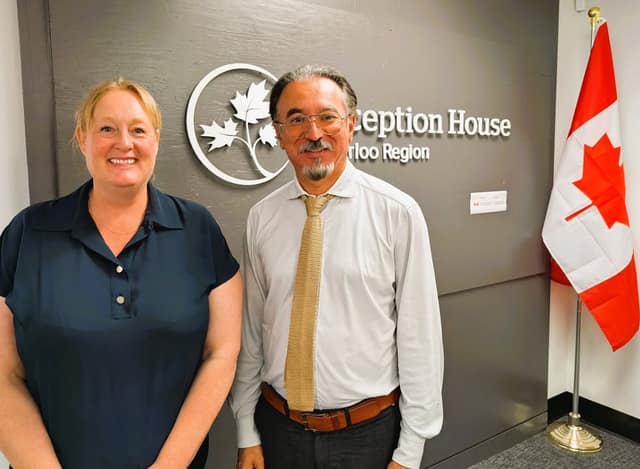 A smiling woman in a navy shirt and a man with glasses, a goatee and a knit tie stand before the Reception House sign and a Canadian flag.
