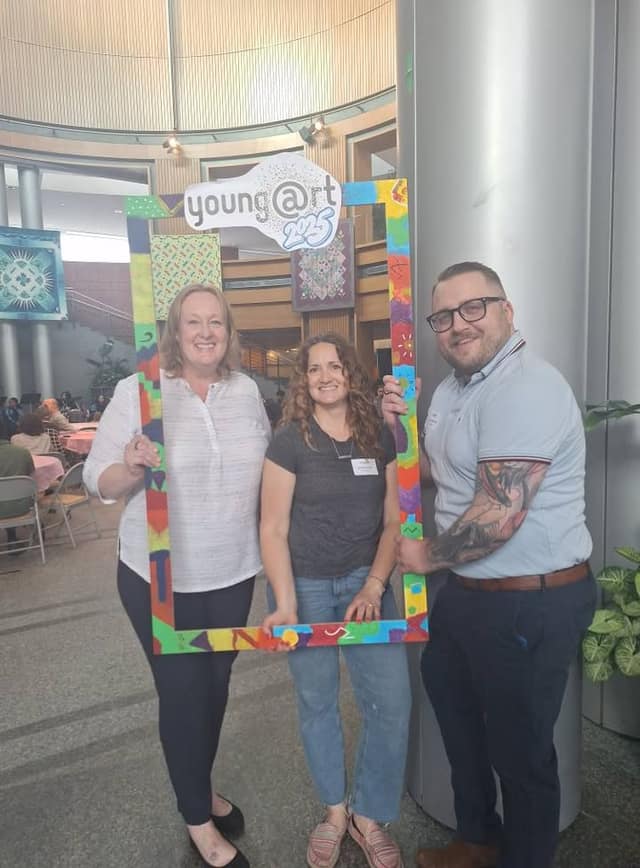 Three smiling people stand holding a colourful painted frame topped with a "young@rt 2025" sign in a bright atrium at City Hall.