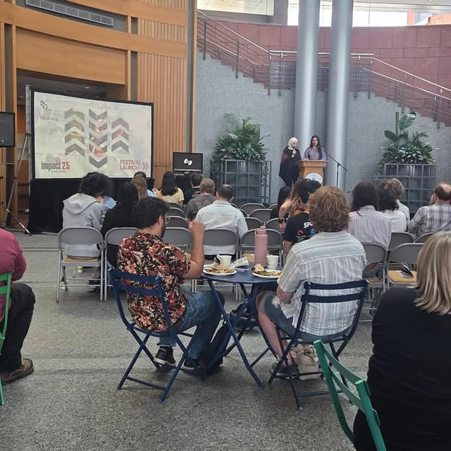 A woman speaks at a wooden podium in a City Hall atrium while seated attendees face a screen reading IMPACT 25 Festival Launch.