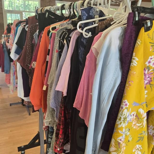 A rack of colourful clothing on hangers, including an orange top, a light blue shirt and a yellow floral blouse, in a hall with a wood floor.
