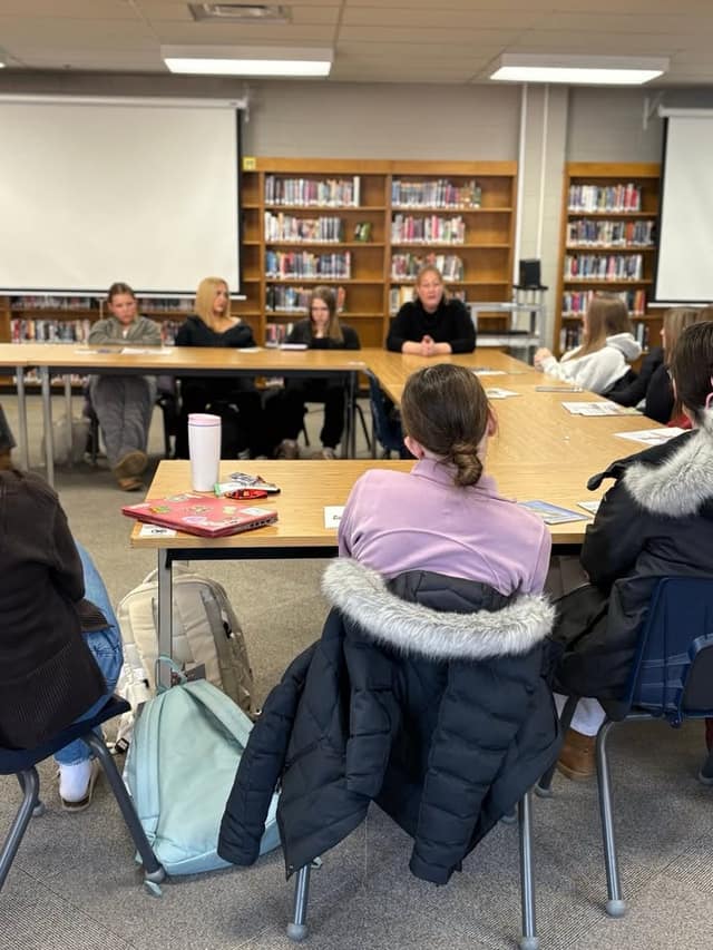 A woman in black sits at the head of a U-shaped table in a school library with students seated around it, jackets on chairs.