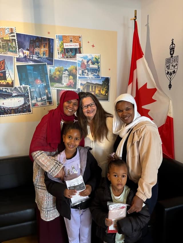 A woman with glasses stands with two women and two young girls in an office beside a Canadian flag and a wall mural of local landmarks.