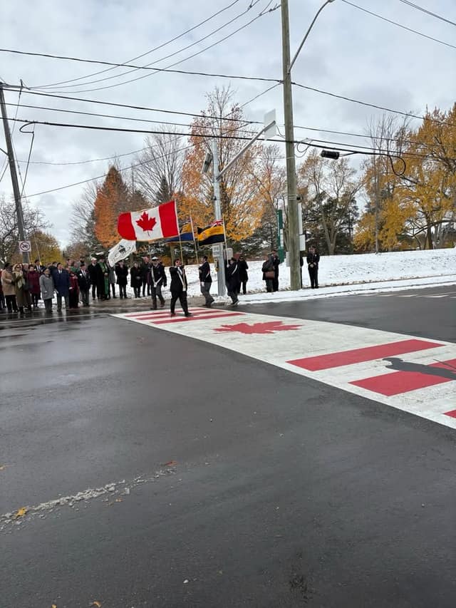 A color party carrying flags walks toward the new crosswalk as onlookers stand in the snow beside autumn trees.