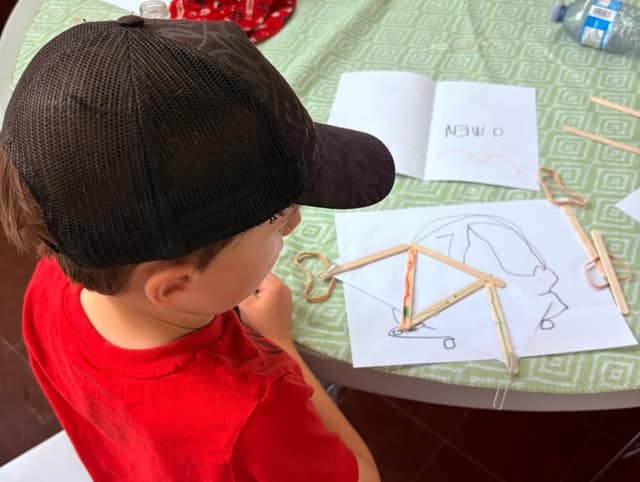 A boy in a black mesh cap and red shirt builds a triangle structure from craft sticks and rubber bands on a table with drawings.