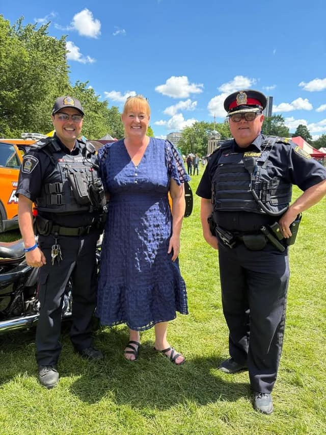 A smiling woman in a blue dress stands on a grassy field between two uniformed police officers, one beside a motorcycle.