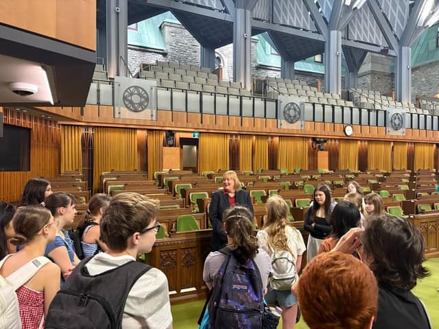 A woman in a dark blazer speaks to a group of students in the House of Commons chamber, with the galleries behind her.