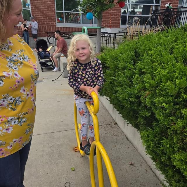 A smiling young girl with blonde curly hair holds a yellow metal bike rack beside a hedge, with people seated behind her.