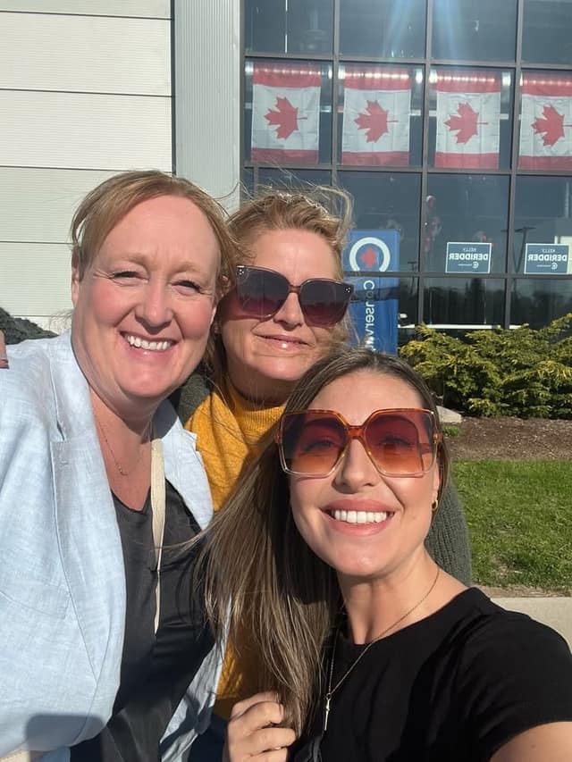 Three smiling women take a selfie outdoors in sunshine in front of windows with Canadian flags and Kelly DeRidder signs.