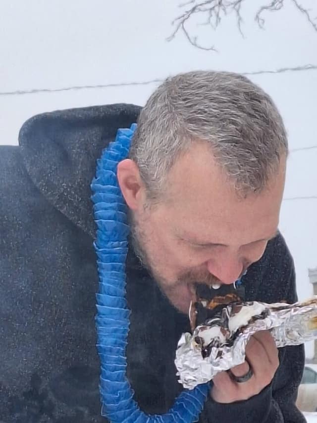 A man with graying hair and a blue lei bites into a foil-wrapped s'more outdoors in falling snow.