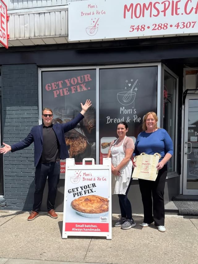 A man with arms outstretched, a woman in an apron and a woman holding a tote stand outside Mom's Bread & Pie Co. beside a pie sign.