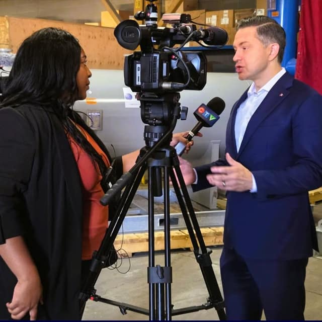 A man in a navy suit speaks to a woman holding a microphone beside a video camera on a tripod in a warehouse.