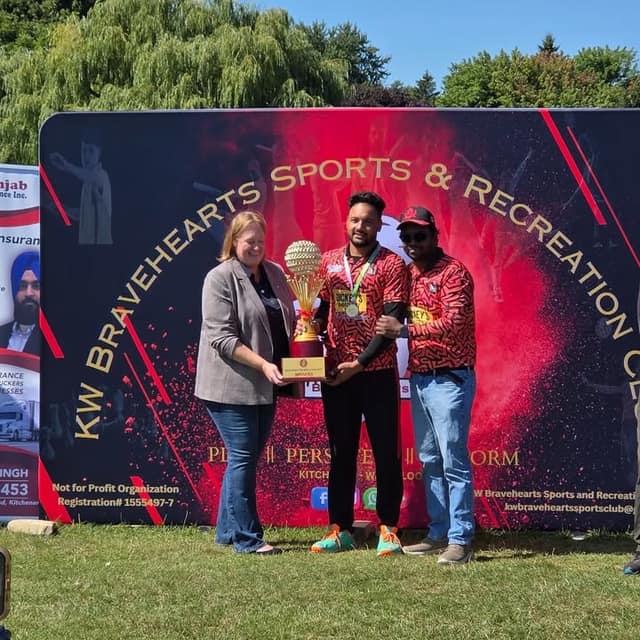 A woman in a gray blazer holds a trophy with a man in a red jersey and medal, and another man, before the Bravehearts banner.