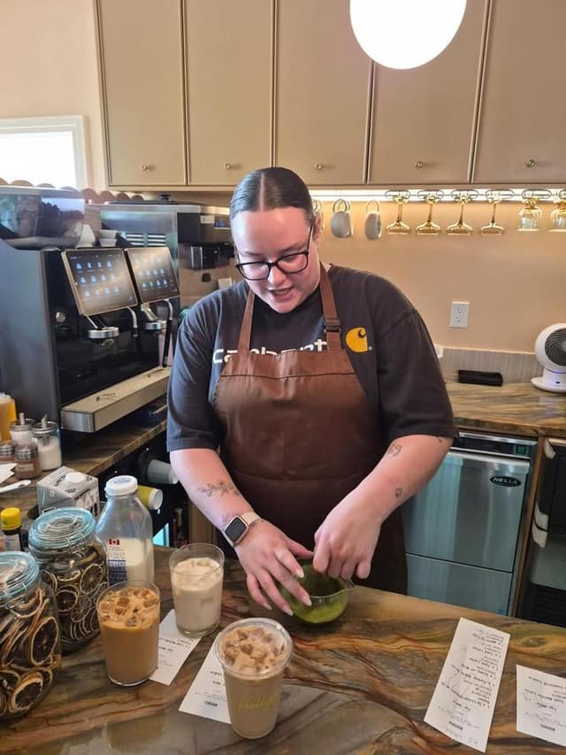A barista in glasses and a brown apron whisks matcha at a counter beside two iced coffees and an espresso machine.