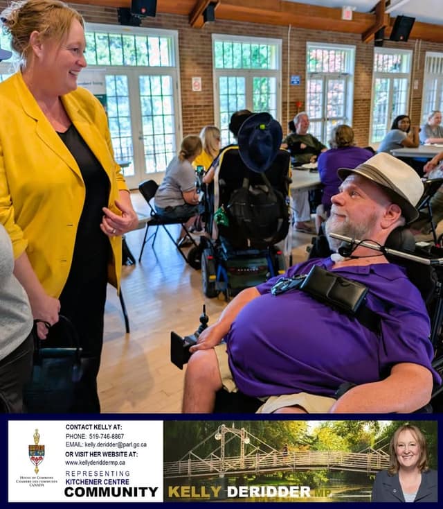 A woman in a yellow blazer chats with a man in a purple shirt and hat using a power wheelchair, with guests seated behind.
