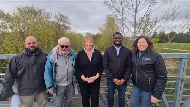 Five people stand smiling in a row on a metal bridge with railings, with budding trees and a grassy field behind them under a cloudy sky.