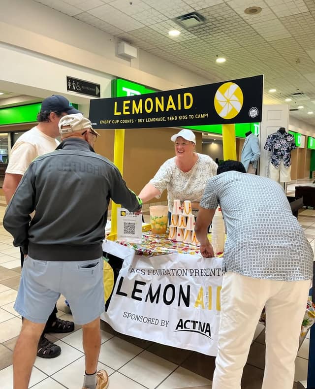 A woman in a white cap smiles behind the LemonAID stand as two men lean in to buy lemonade; paper cups are stacked on the table.