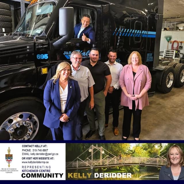 A man in a suit gives a thumbs up from a black transport truck cab while five people pose in front of it in a garage.