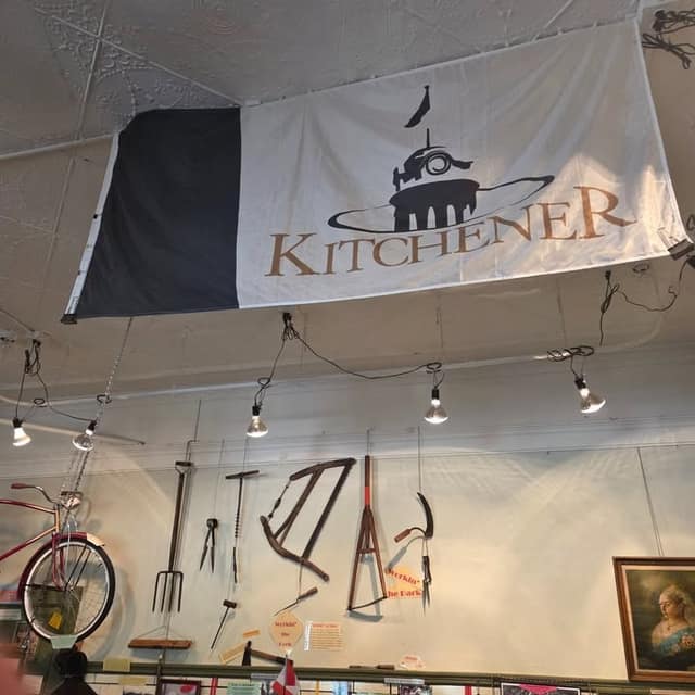 Large Kitchener flag with a clock tower hangs from the ceiling above antique hand tools, a red bicycle and a framed portrait.